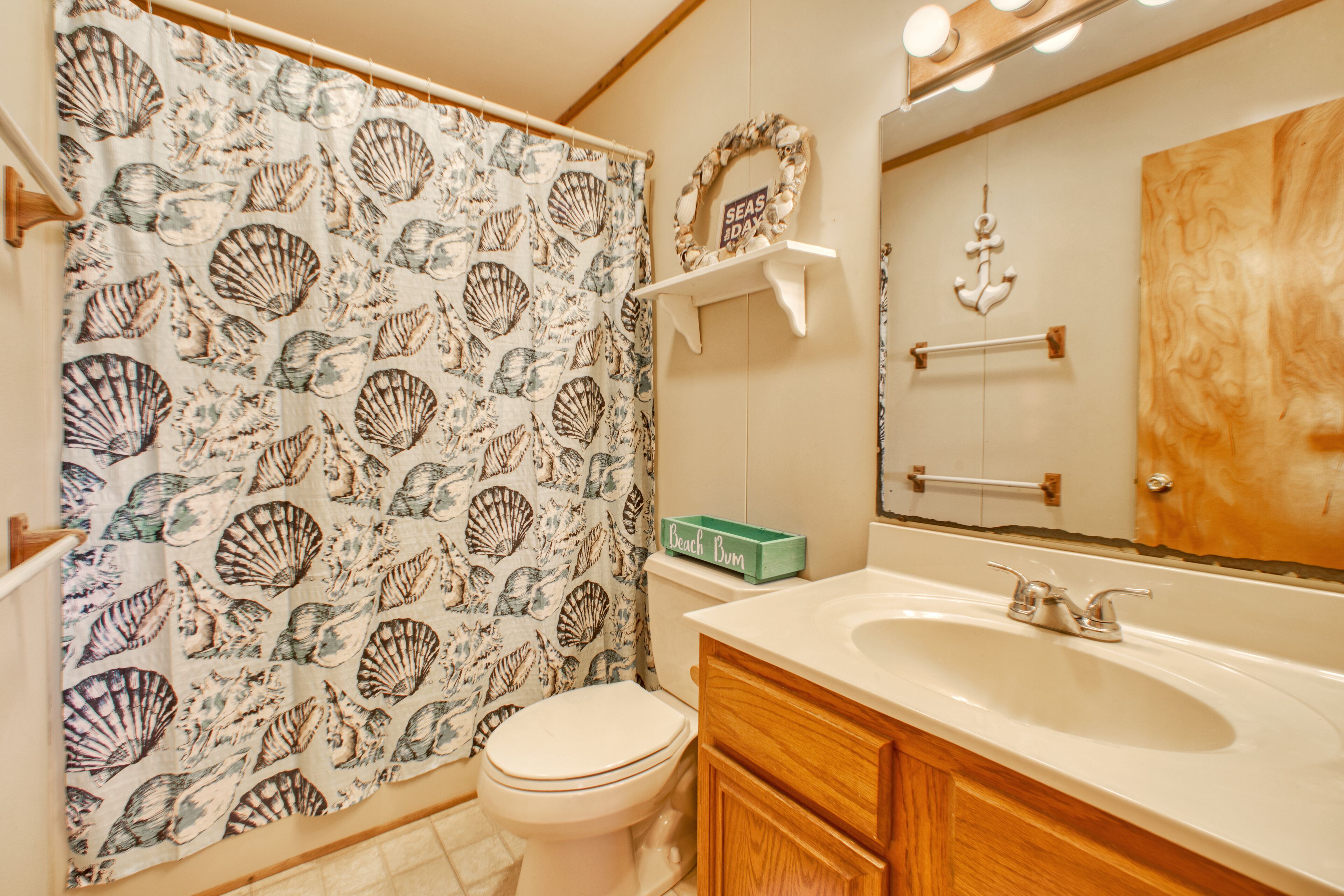 Une salle de bain équipée de toilettes, d’un lavabo et d’un rideau de douche orné d’un motif de coquillages. On y trouve des meubles en bois et un miroir au-dessus du lavabo.