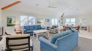 A spacious living room with a large blue sofa, a white coffee table, and a ceiling fan. There is a dining area with a table and chairs in the background. - Gray Haven #11-DE: Oceanfront, dog-friendly, private hot tub, pool access (Avon)