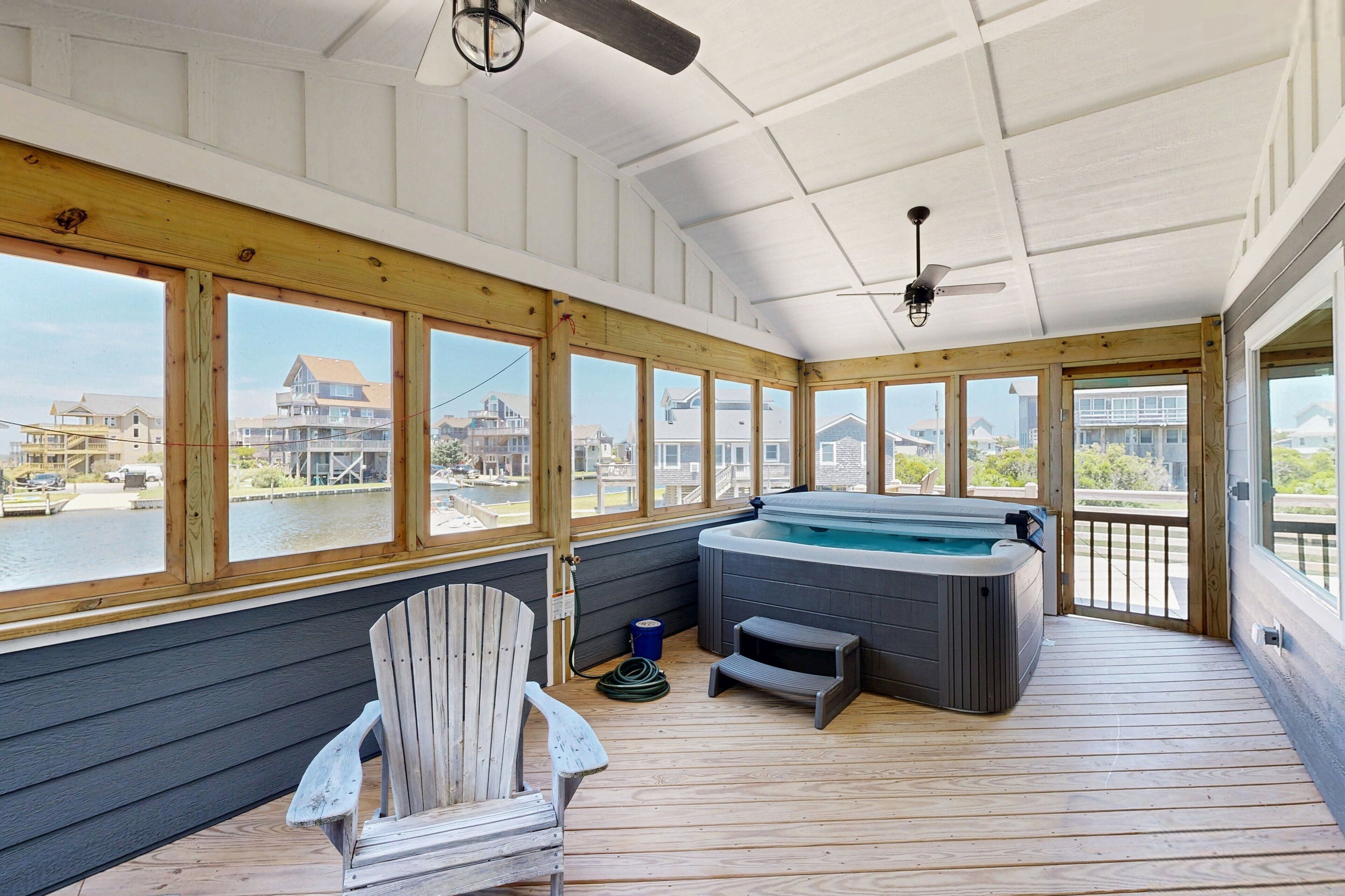 A covered outdoor deck with a hot tub, wooden chairs, and large windows overlooking a body of water.