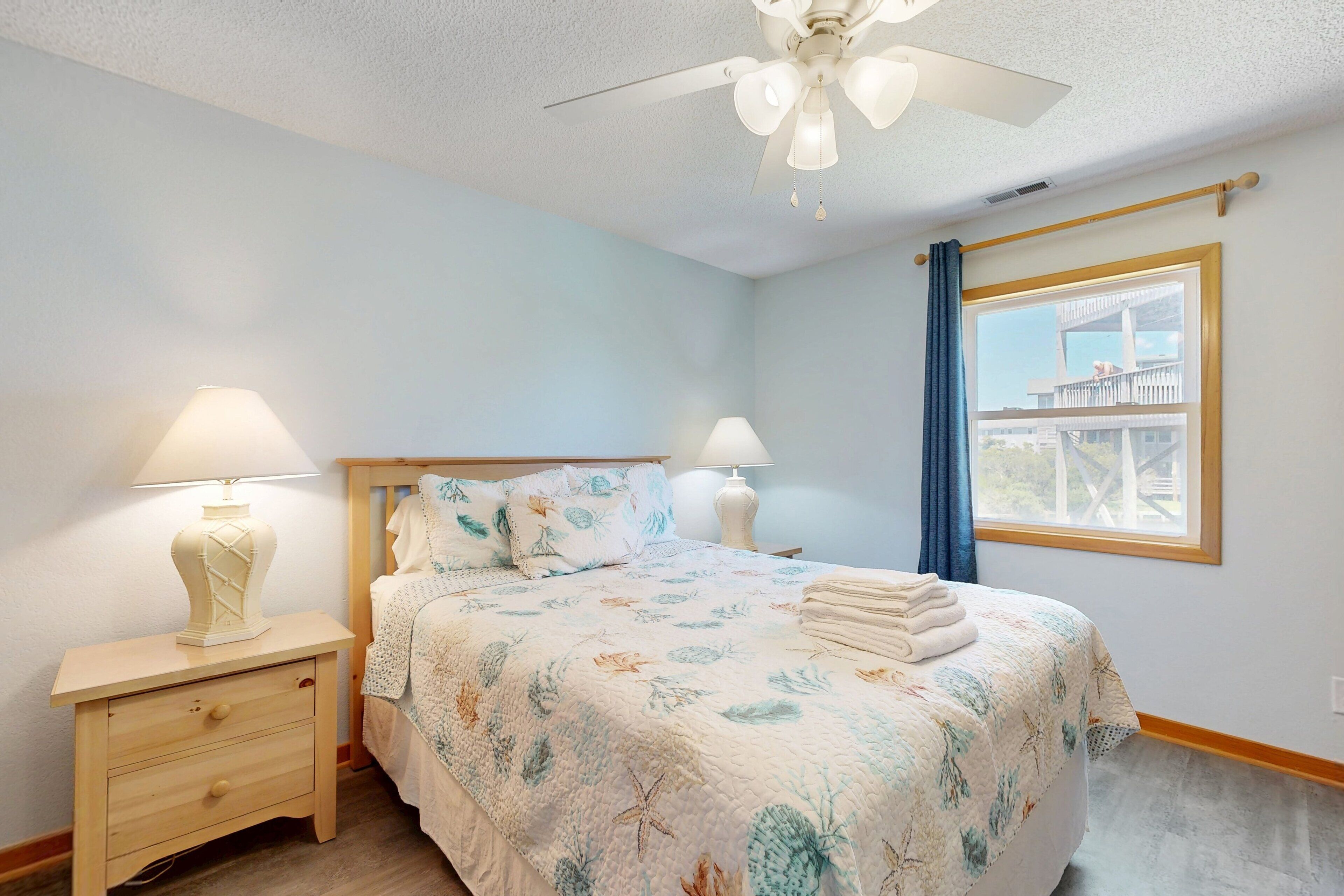 A bedroom with a bed, two bedside lamps, a ceiling fan, a window with a view, and a wooden nightstand with drawers.