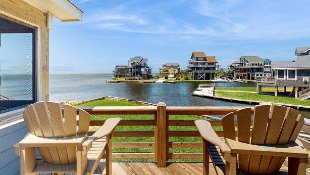 A deck with two wooden chairs and a wooden railing overlooking a water body with buildings in the background. - Waterfront Home on Bulk-Headed Lot w/ Private Hot Tub, Sound View, Shared Pool (Avon)