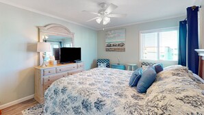 Een slaapkamer met een bed, een televisie, een dressoir, een plafondventilator, een raam met gordijnen en een schilderij aan de muur.