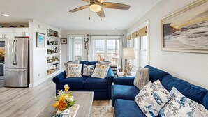 Un salon moderne avec un canapé bleu, un ventilateur de plafond et une cuisine en arrière-plan.