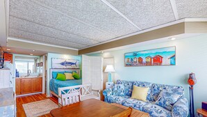 A cozy living area with a wooden dining table, a blue patterned sofa, and a painting of houses on the wall. - Sea Alcove: Colorful studio near the ocean with pool & kitchenette (Atlantic Beach)