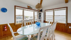 Een eetruimte met een glazen tafel, witte stoelen, een blauwe kom en uitzicht op zee door grote ramen.