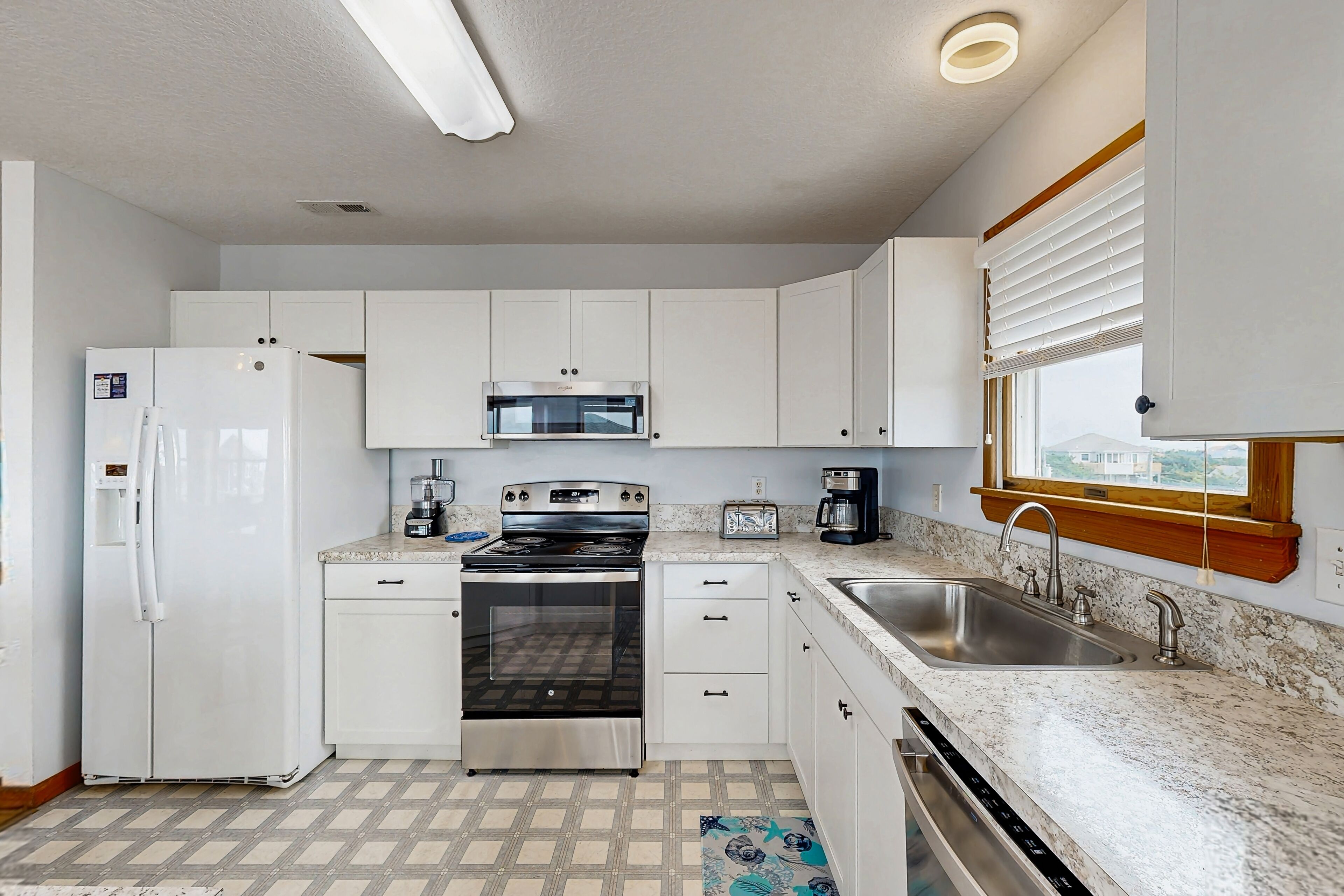 Cocina compacta con armarios blancos, refrigerador de acero inoxidable, horno empotrado, microondas, tostadora y lavaplatos con ventana arriba.