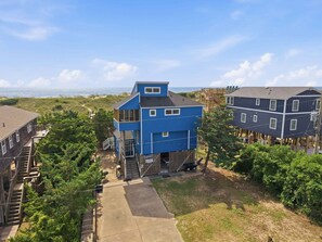 A coastal residential area with a blue house featuring a front porch and a deck, surrounded by greenery and a clear view of the ocean.
