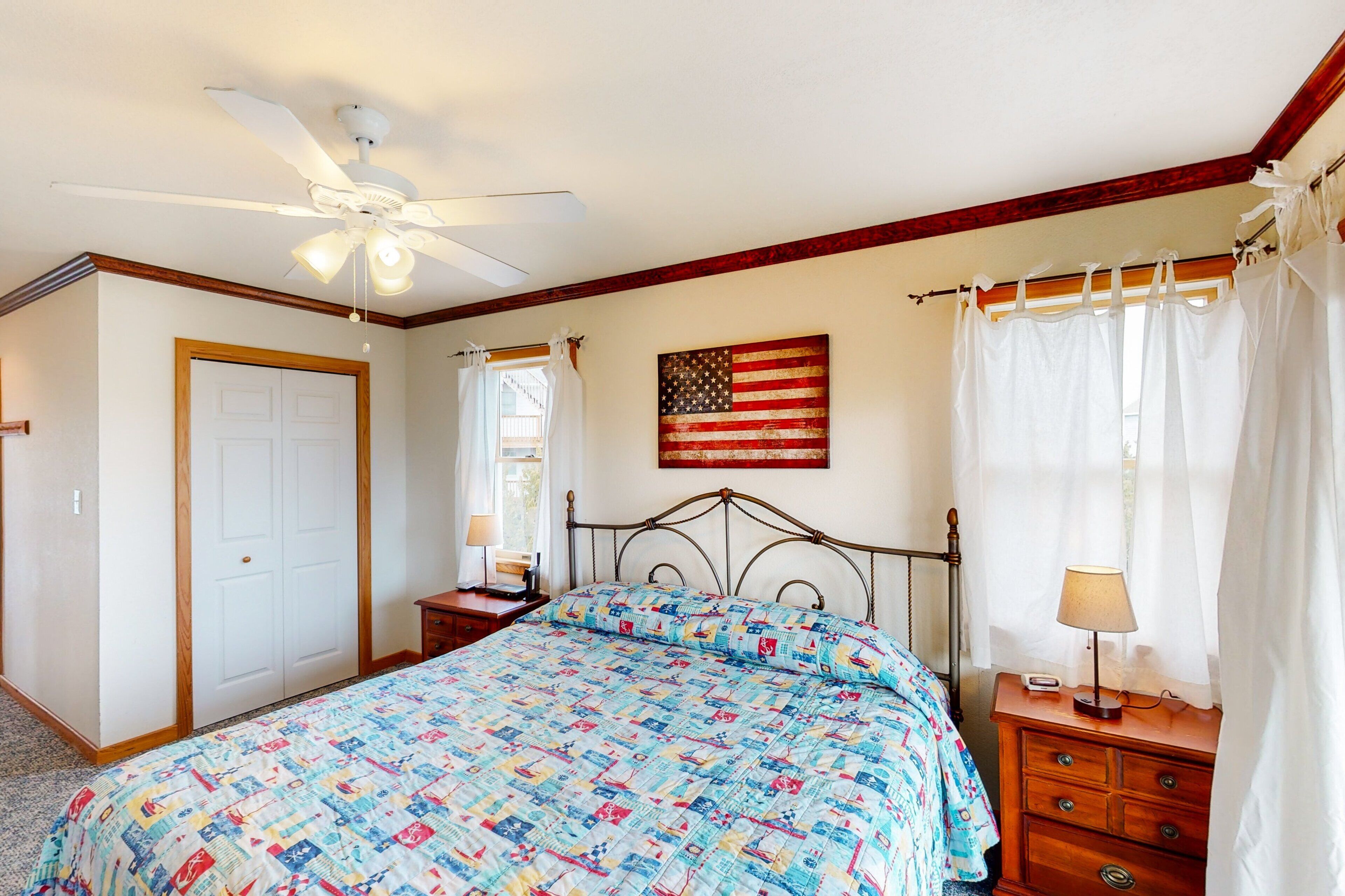 A bedroom with a bed, a ceiling fan, a nightstand with a lamp, a dresser, a window with curtains, and an American flag on the wall.