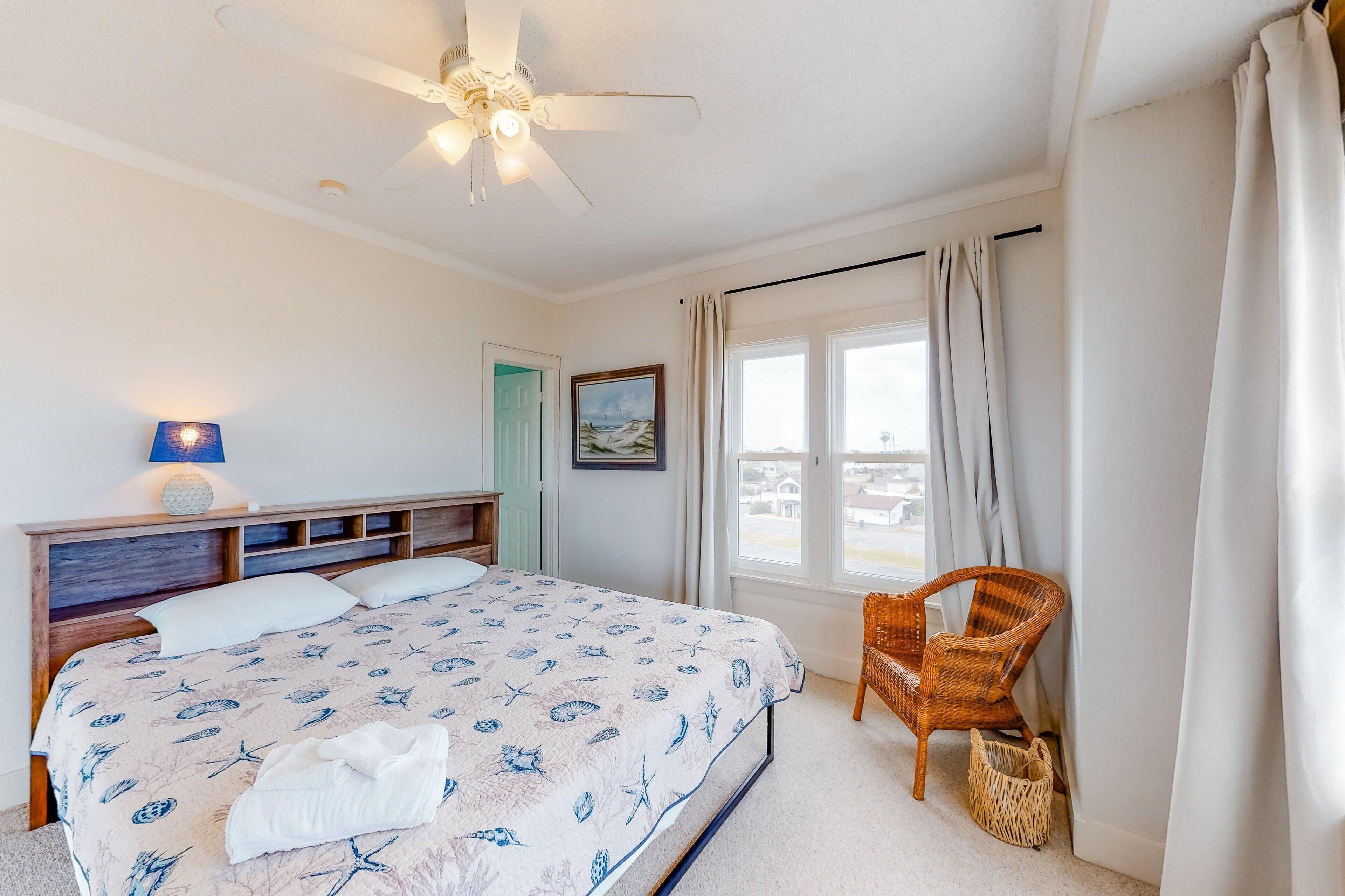 Une chambre à coucher avec un lit, un ventilateur de plafond, une fenêtre avec des rideaux, un fauteuil en osier et un petit panier.