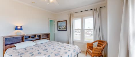 Un dormitorio con cama, ventilador de techo, ventana con cortinas, una silla de mimbre y una cesta pequeña.