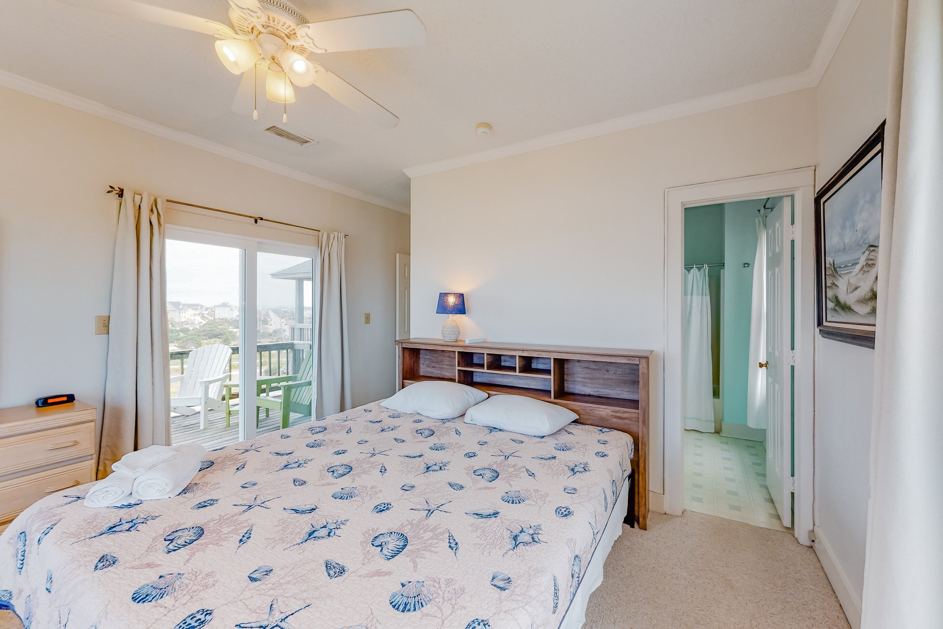 Un dormitorio con cama, mesita de noche, ventilador de techo, ventana con cortinas y puerta que da a otra habitación.