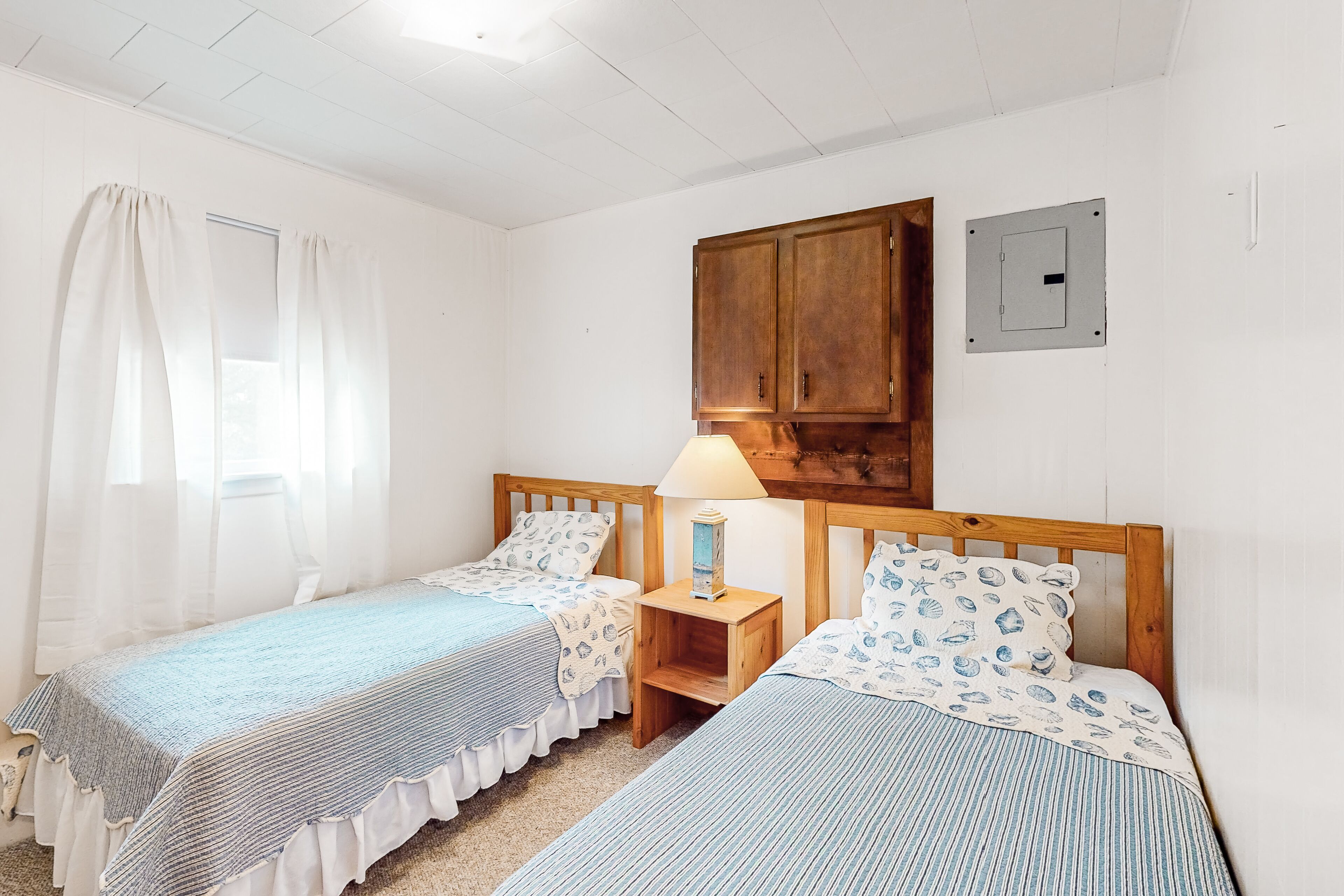 A bedroom with two single beds, a wooden wardrobe, a nightstand with a lamp, and a window with sheer curtains.