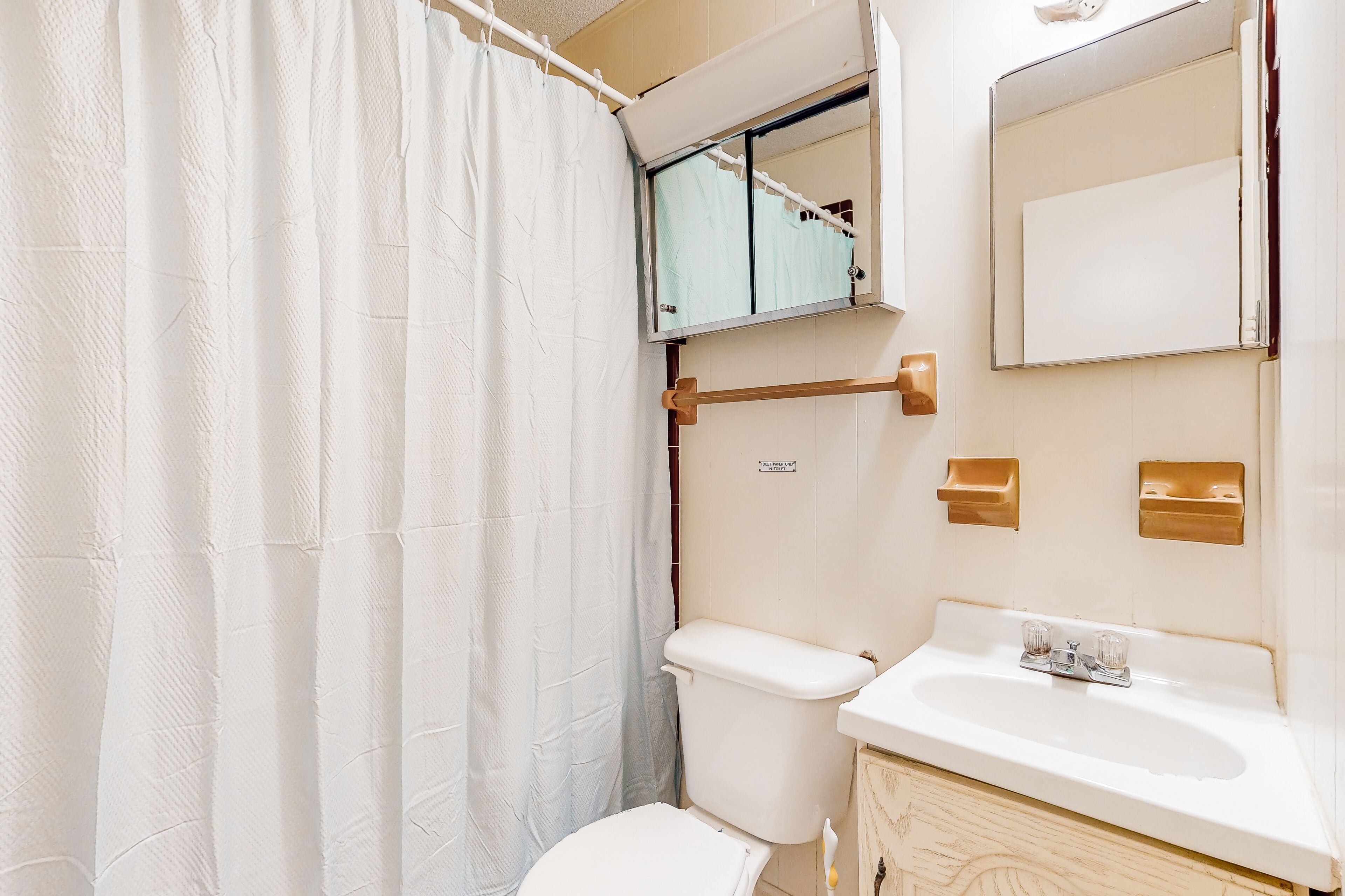 Une salle de bain avec des toilettes blanches, un lavabo, un miroir et un rideau de douche.