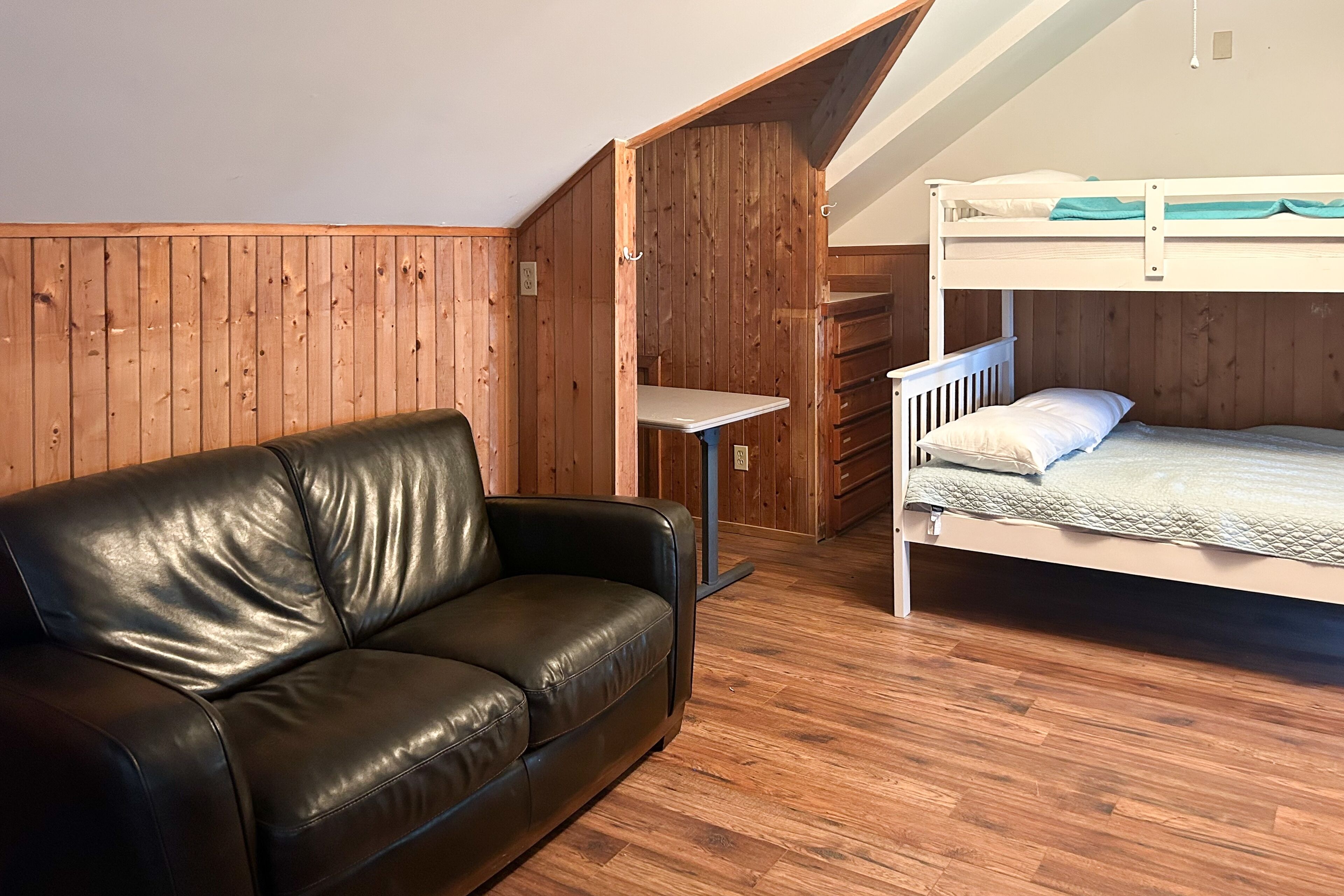 A room with a wooden floor, a black leather sofa, a bunk bed, and a desk.