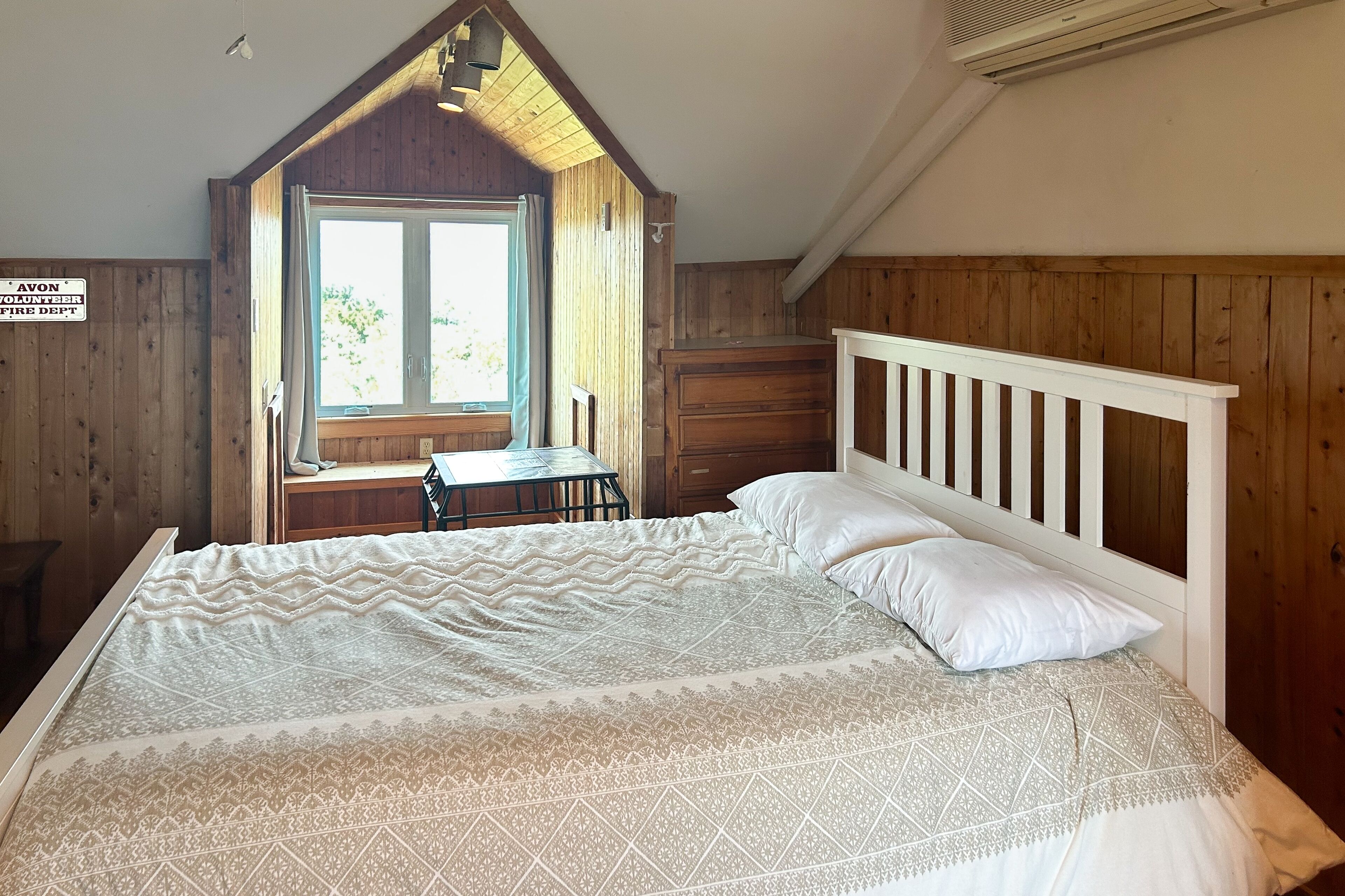 A bedroom with a wooden sloped ceiling, a bed with white linens, a small table, and a window with curtains.