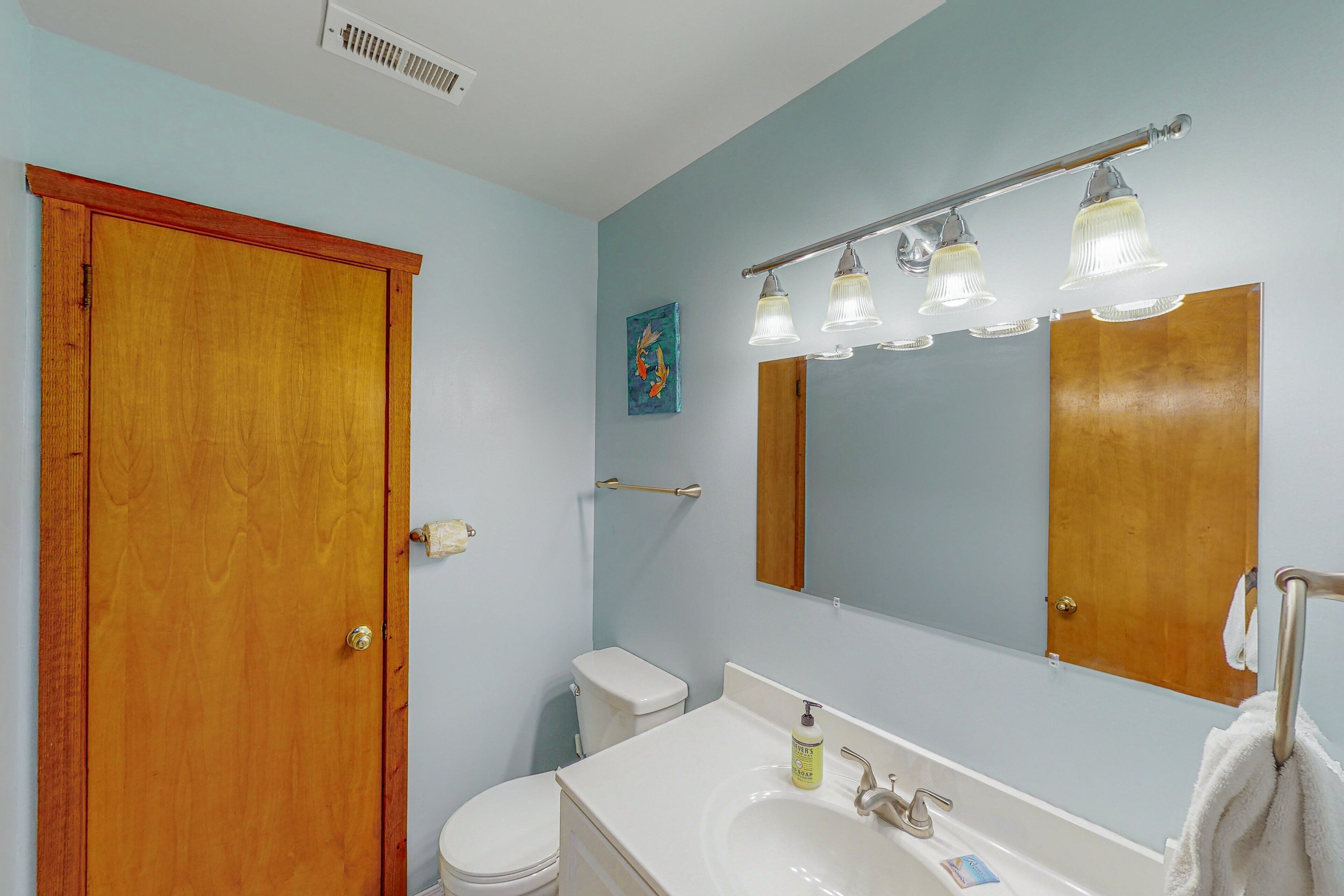 Un baño con puerta de madera, inodoro blanco, lavabo con espejo y toalla colgada en la pared.