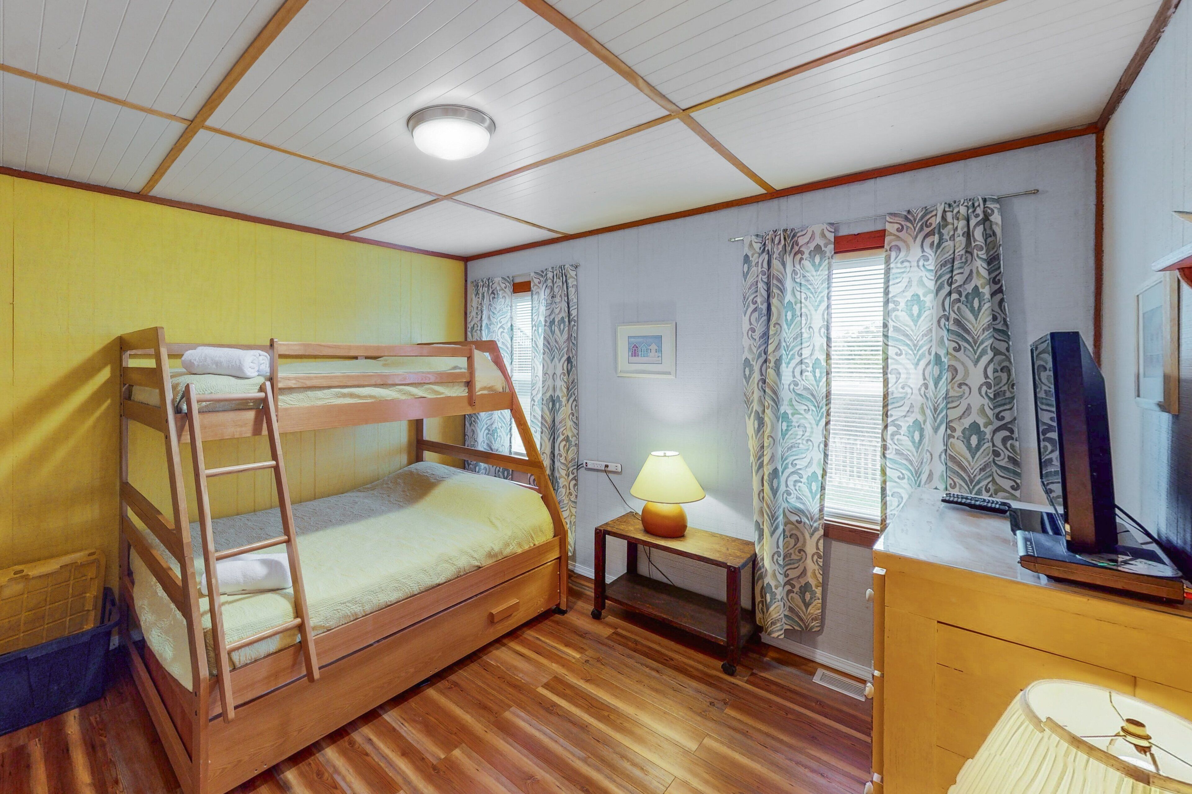 A bunk bed room with a wooden floor, a desk with a lamp, a television, and patterned curtains.