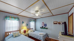 A room with two single beds, a ceiling fan, a TV on a stand, and a wall-mounted shelf with a decorative item. - Sand Dollar #13-HC: Oceanfront, Dog-Friendly, Private Hot Tub (Avon)
