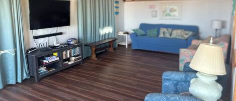Una sala de estar con televisor de pantalla plana, una estantería, un sofá azul y un sillón estampado.