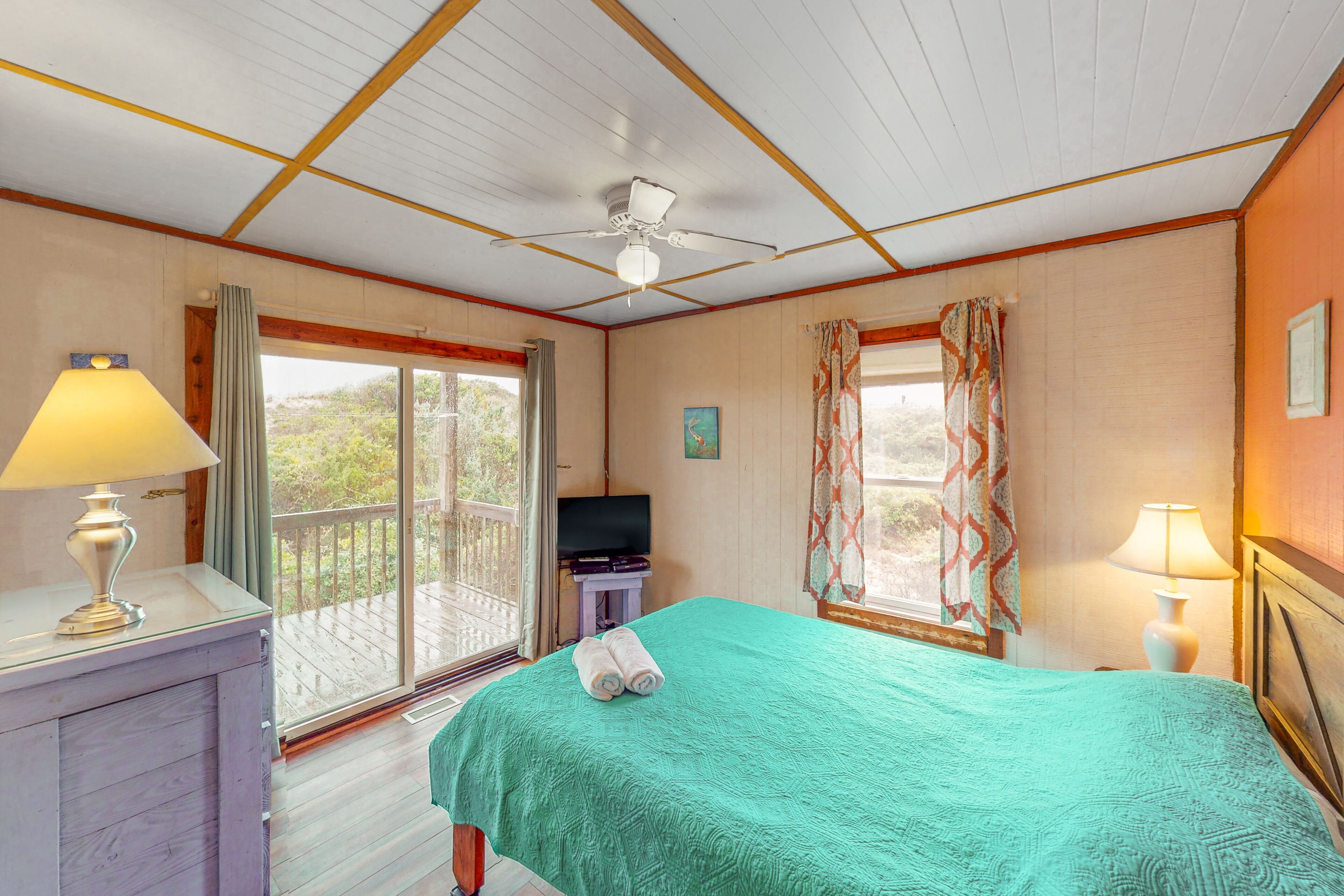 A room with a bed, a television, a ceiling fan, and a balcony with curtains.