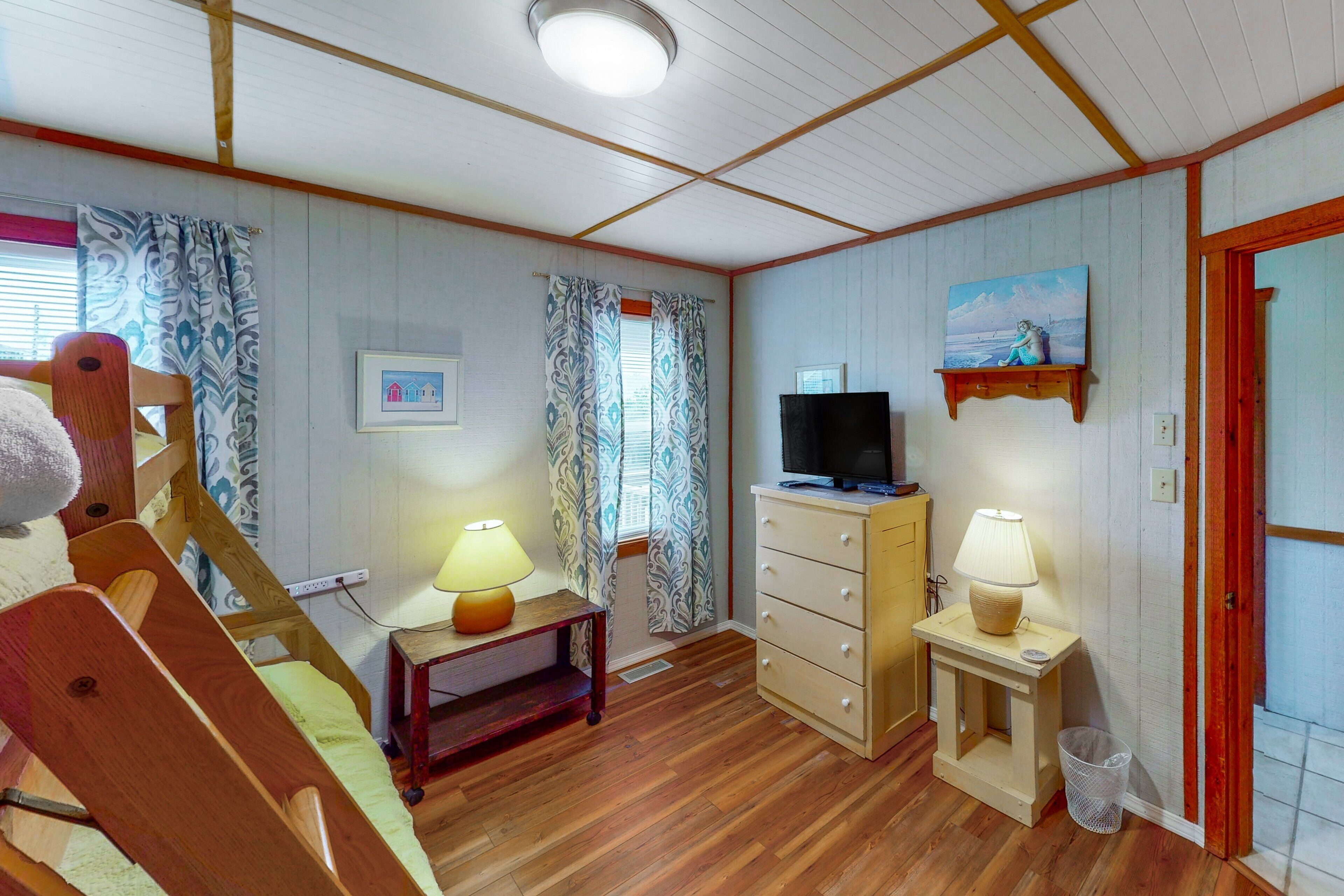Ein gemütliches Zimmer mit Holzboden, einem Fernseher auf einem Schrank, einem kleinen Tisch mit Lampe, einem Sessel und einer Tür mit rotem Rahmen.