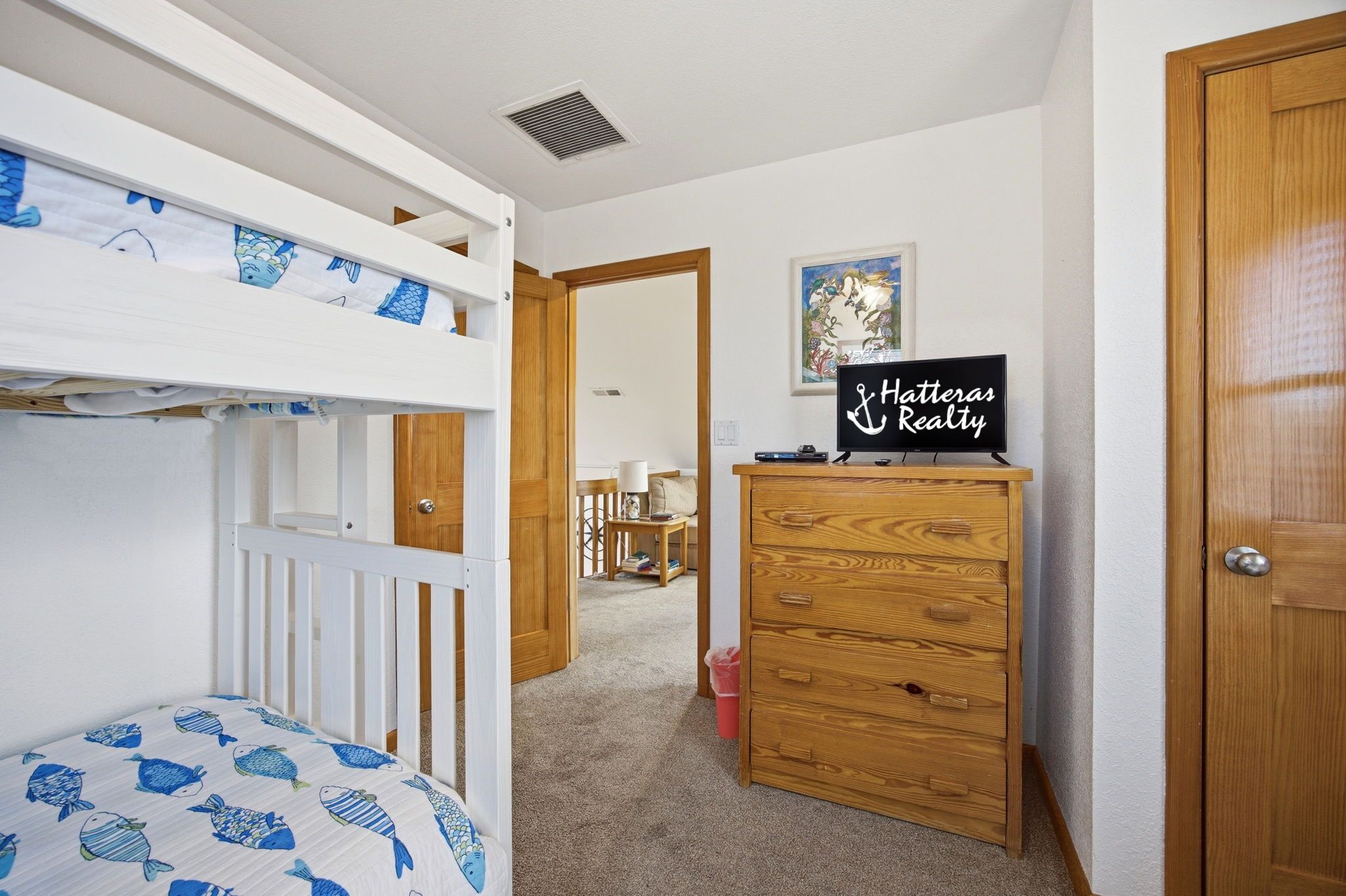 A bedroom with a bunk bed, a wooden dresser, a desk with a sign, and a door leading to another room.