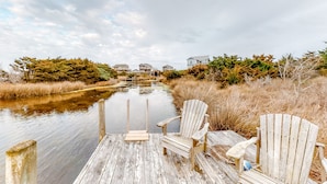 Un muelle de madera con dos sillas Adirondack mirando un río tranquilo, rodeado de pastos altos y una fila de casas al fondo.