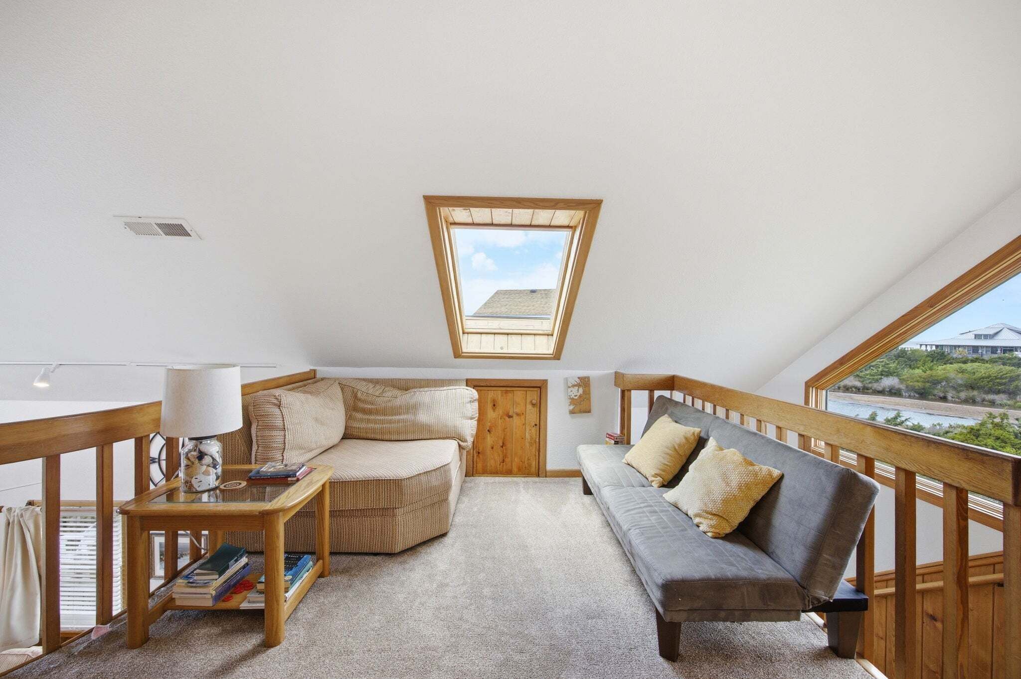 A loft bedroom with a sloped ceiling, a skylight, a wooden railing, a sofa, a chair, and a small table with books.