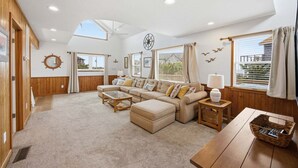 Amplio salón con un sofá grande, una mesa de centro y una mesa de comedor de madera. Hay ventanas con cortinas y vistas al exterior.