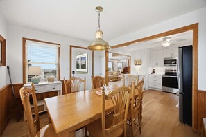 A dining area with a wooden table and chairs, a hanging light fixture, and a view into the kitchen.