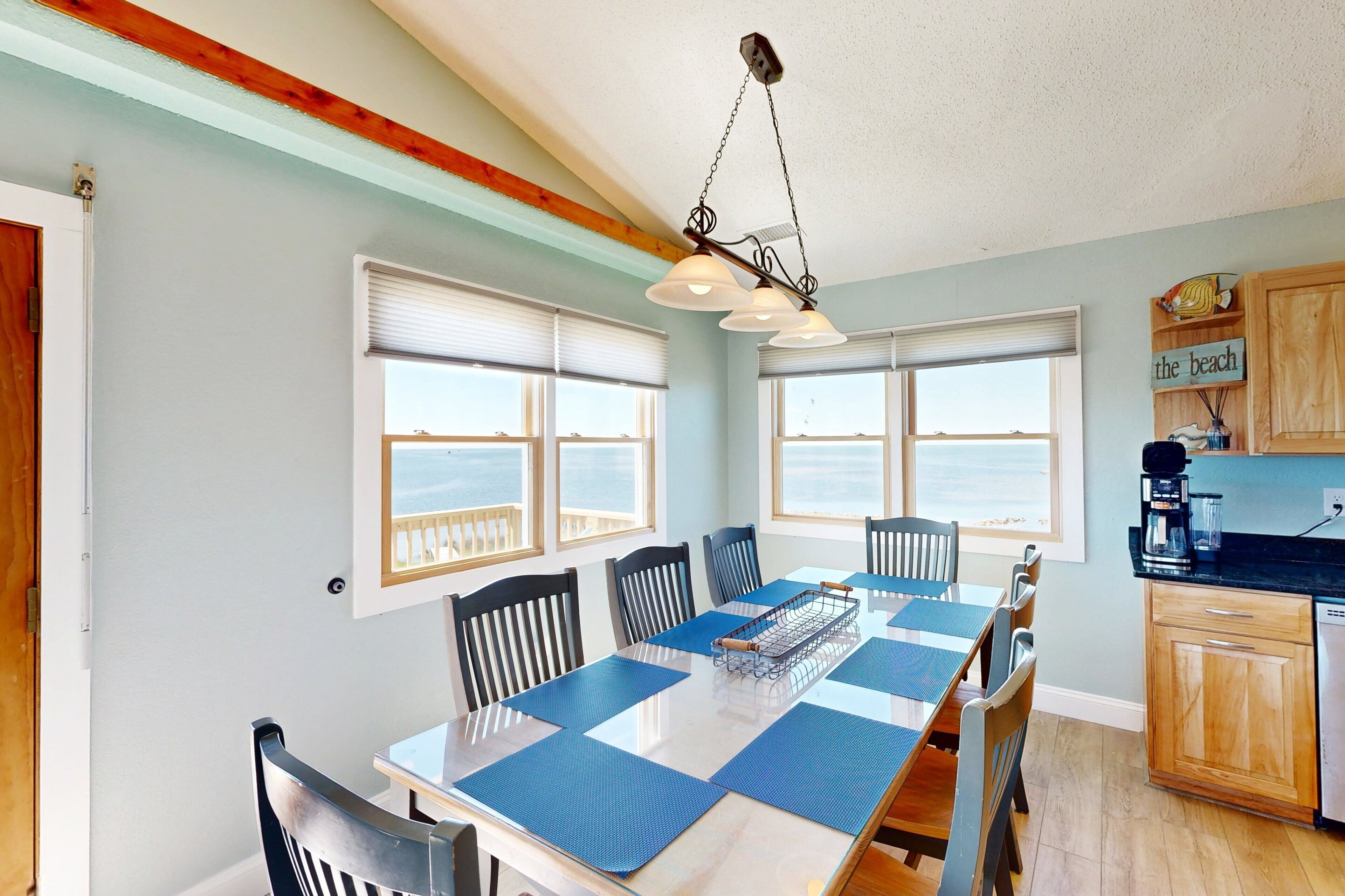 A dining area with a long table, blue placemats, and black chairs. There are windows with blinds, a wooden ceiling, and a kitchen area with wooden cabinets and a coffee maker.