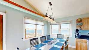 Un comedor con una mesa larga, individuales azules y sillas negras. Se observan ventanas con persianas, techo de madera y una zona de cocina con armarios de madera y una cafetera.