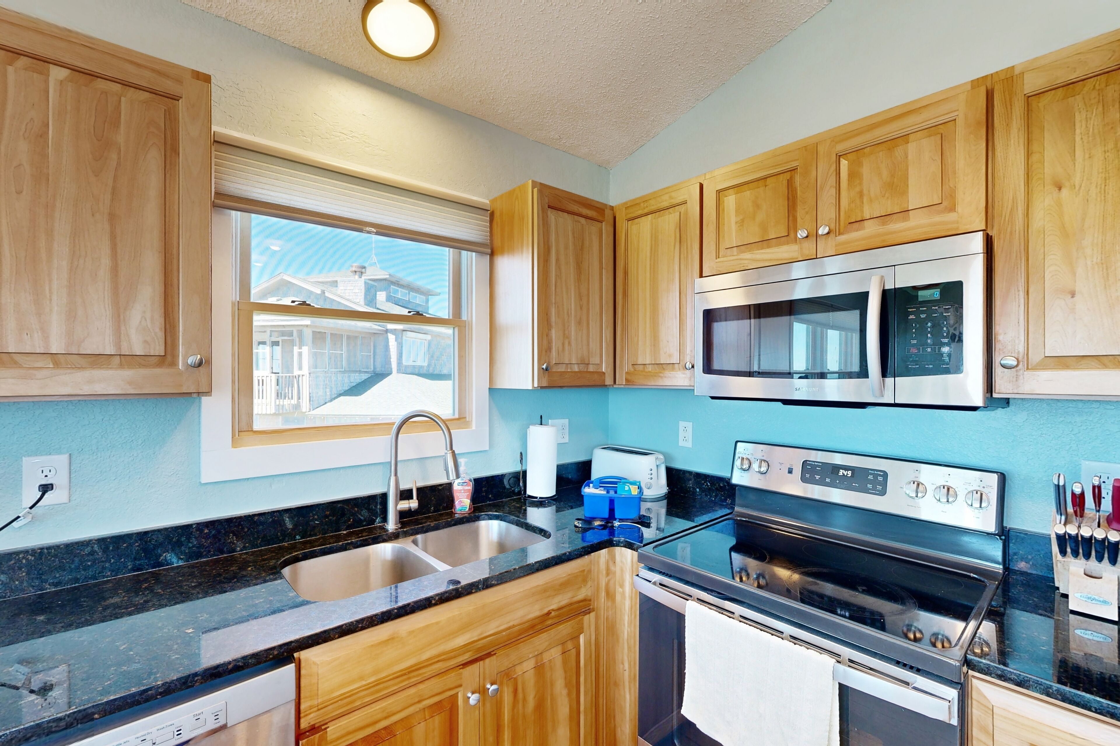Een moderne keuken met houten kastjes, roestvrijstalen apparatuur, een inbouwoven en een raam met uitzicht op een huis.