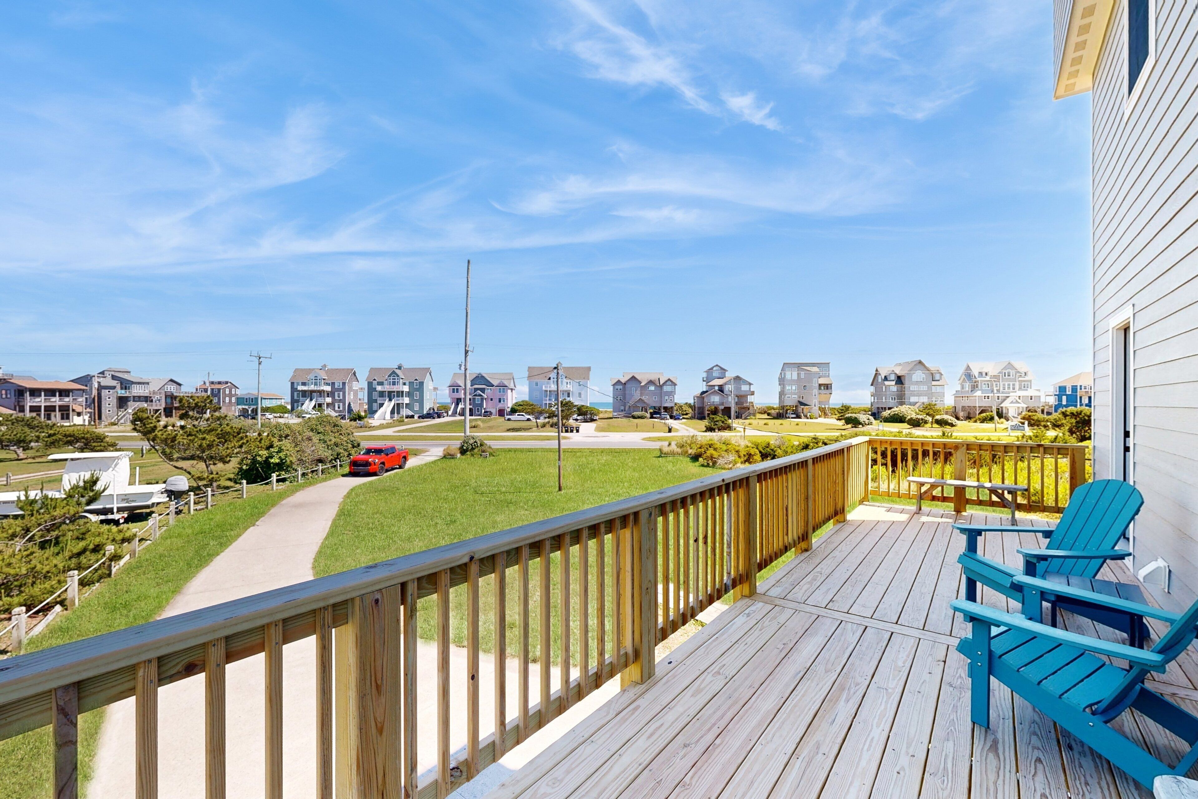 A wooden deck with blue chairs, a clear view of a road with a red car, and a row of houses in the background.