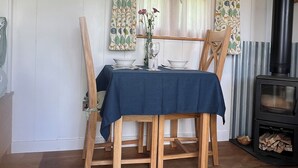 A dining area with a table set for two, a vase with flowers, and a wood-burning stove. - Luxury Shepherd Hut with Private Hot Tub & Views of the Somerset Levels (Langport)