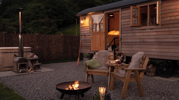 A wooden cabin with a fire pit, outdoor seating, and a view of the forest. - Luxury Shepherd Hut with Private Hot Tub & Views of the Somerset Levels (Langport)