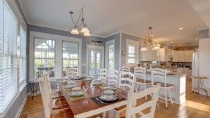 A spacious dining area with a long wooden table set for a meal, surrounded by white chairs. There are large windows providing natural light and a view of the outdoors. - Huge, canal-front home w/private hot tub, WiFi, central AC, washer & dryer, dock (Frisco)