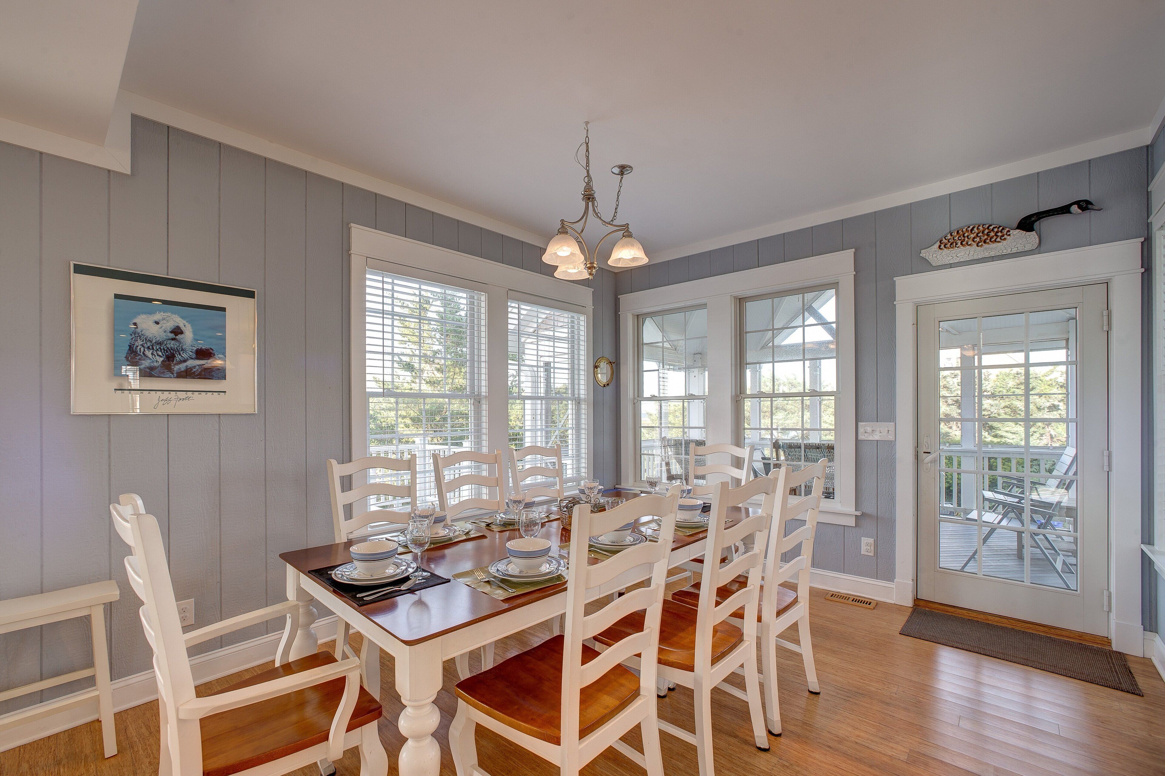 Un coin repas avec une table en bois dressée pour six personnes, des chaises blanches et un lustre.