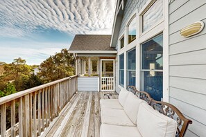 A wooden deck with a white sofa and metal railing, overlooking a residential area with trees and a house.