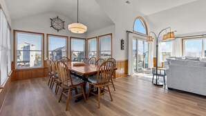 Un vaste espace de salle à manger avec une table et des chaises en bois, de grandes fenêtres équipées de stores et une vue sur la ville.