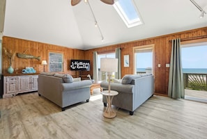 A spacious living room with two sofas, a small round table, and a television. A wooden wall paneling and a large window with a view of the sea are visible.