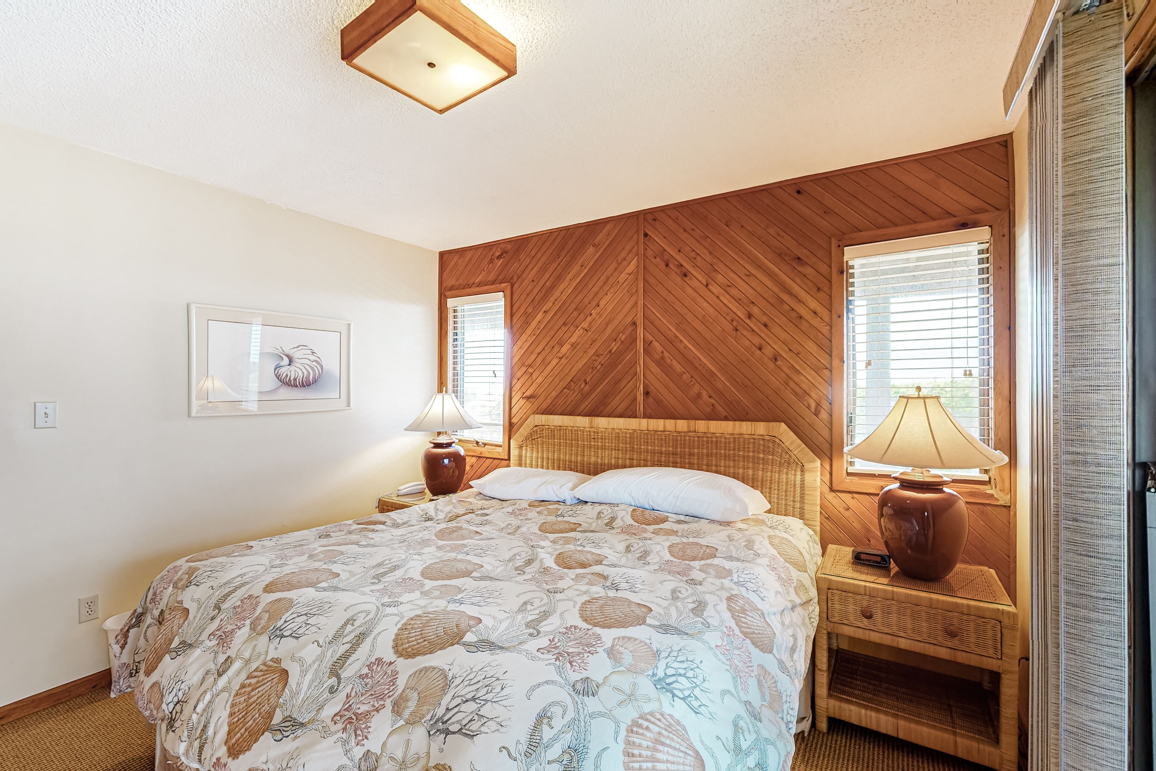 A bedroom with a bed, wooden headboard, two bedside tables with lamps, a framed picture on the wall, and a window with blinds.