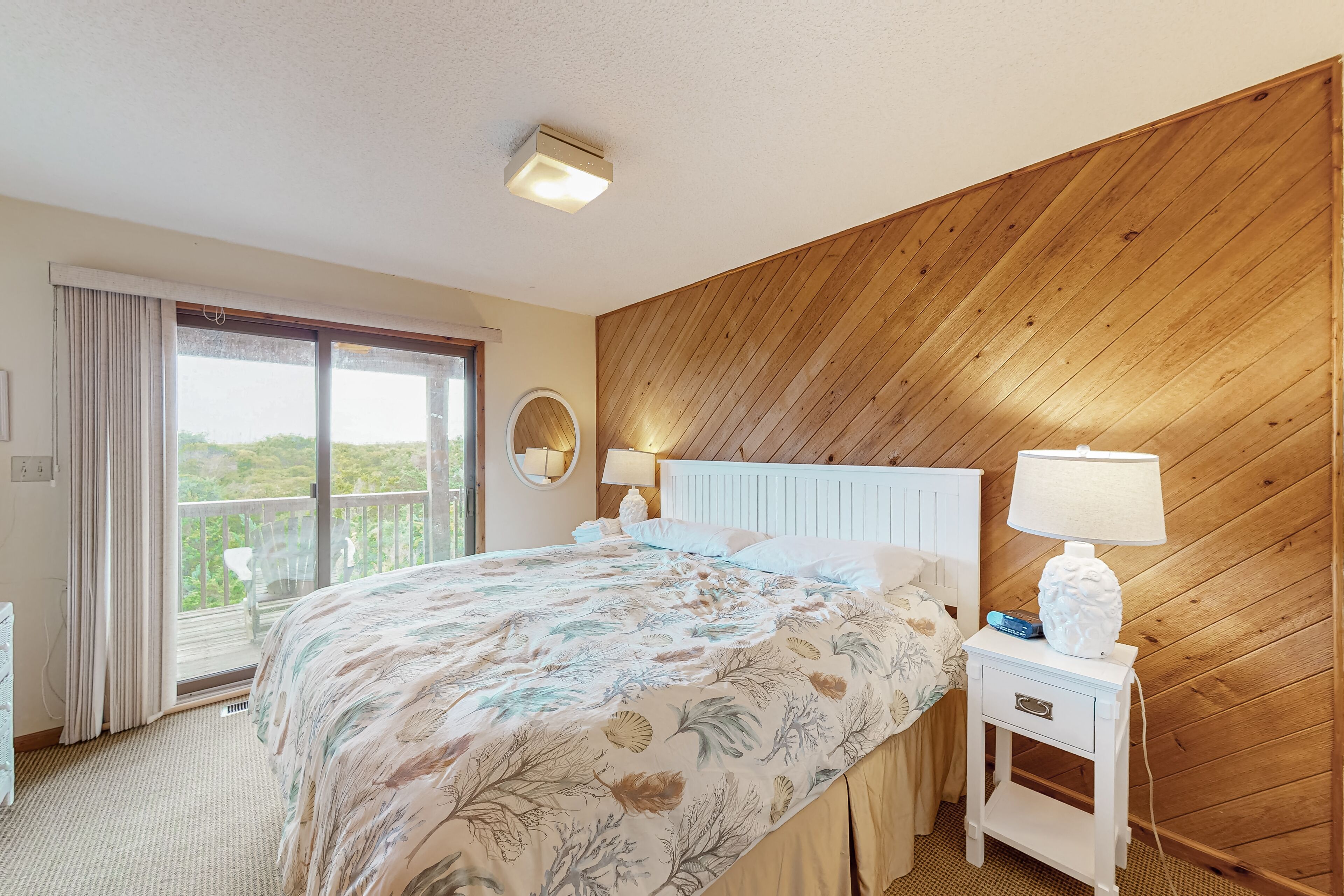 Une chambre avec un grand lit, une table de chevet avec une lampe, une fenêtre avec des rideaux et une vue sur la verdure à l’extérieur.