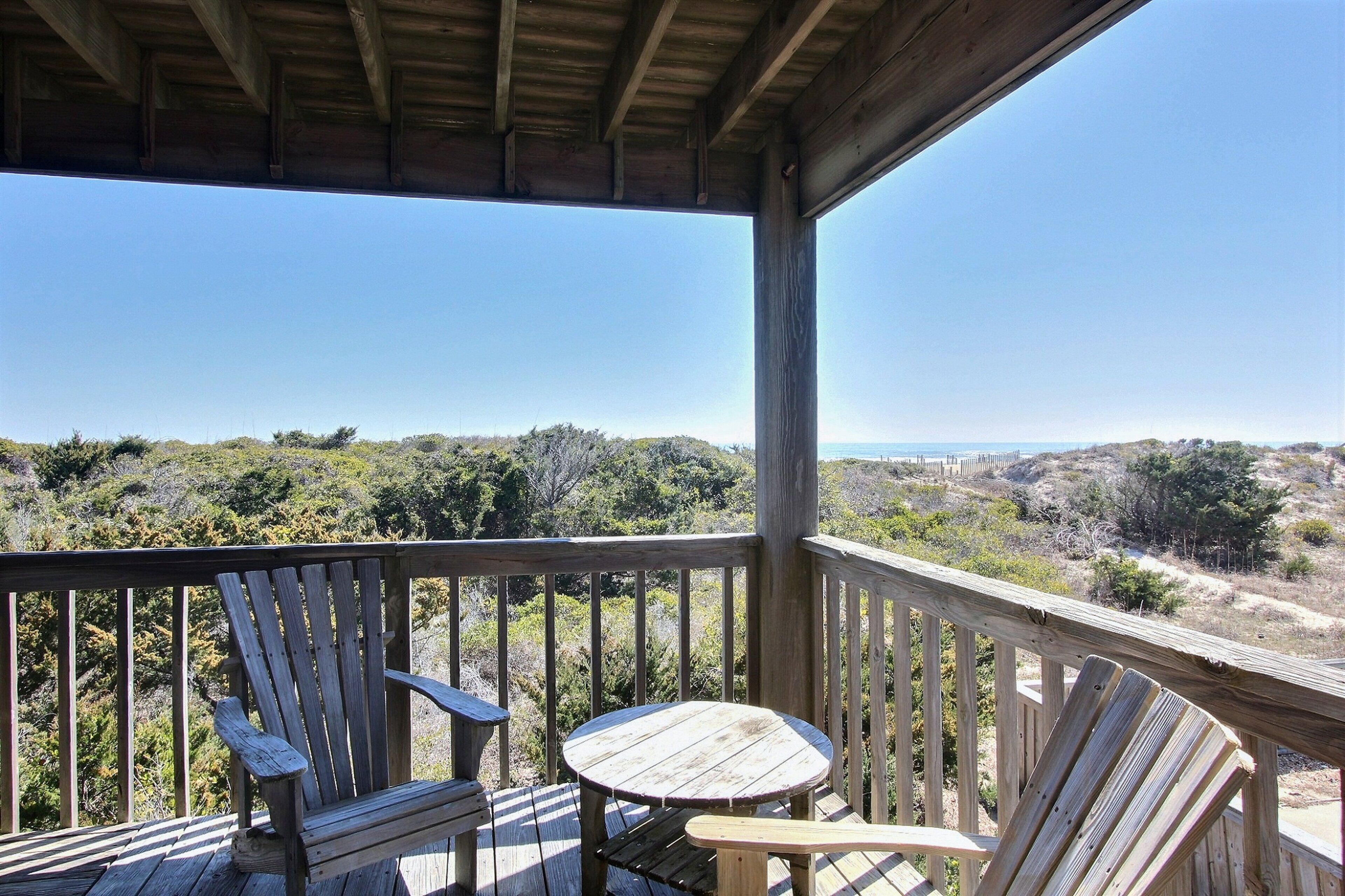 A wooden deck with two Adirondack chairs and a small table, overlooking a natural landscape with trees and hills.