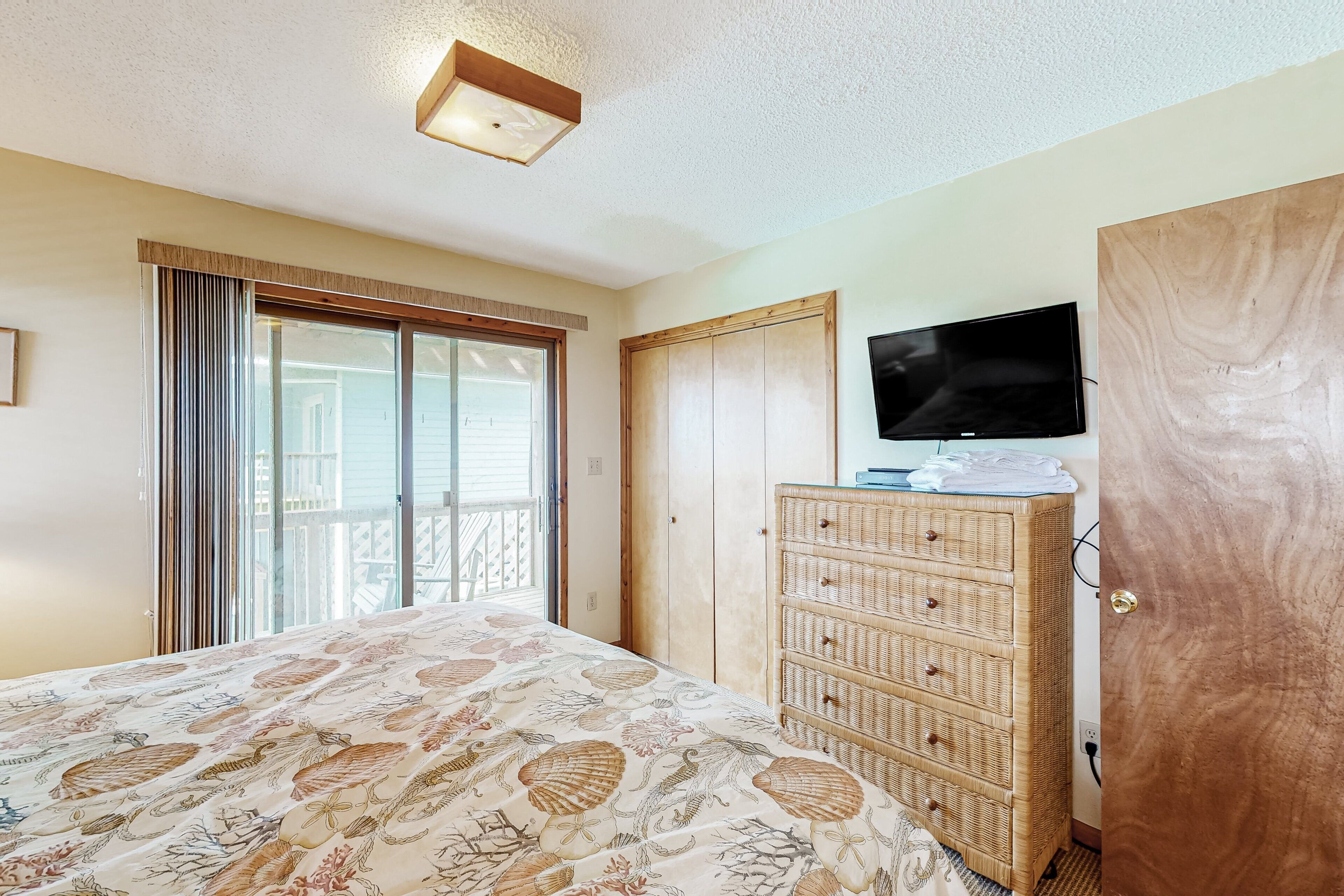 A bedroom with a bed, a dresser, a television, and a sliding glass door to a balcony.