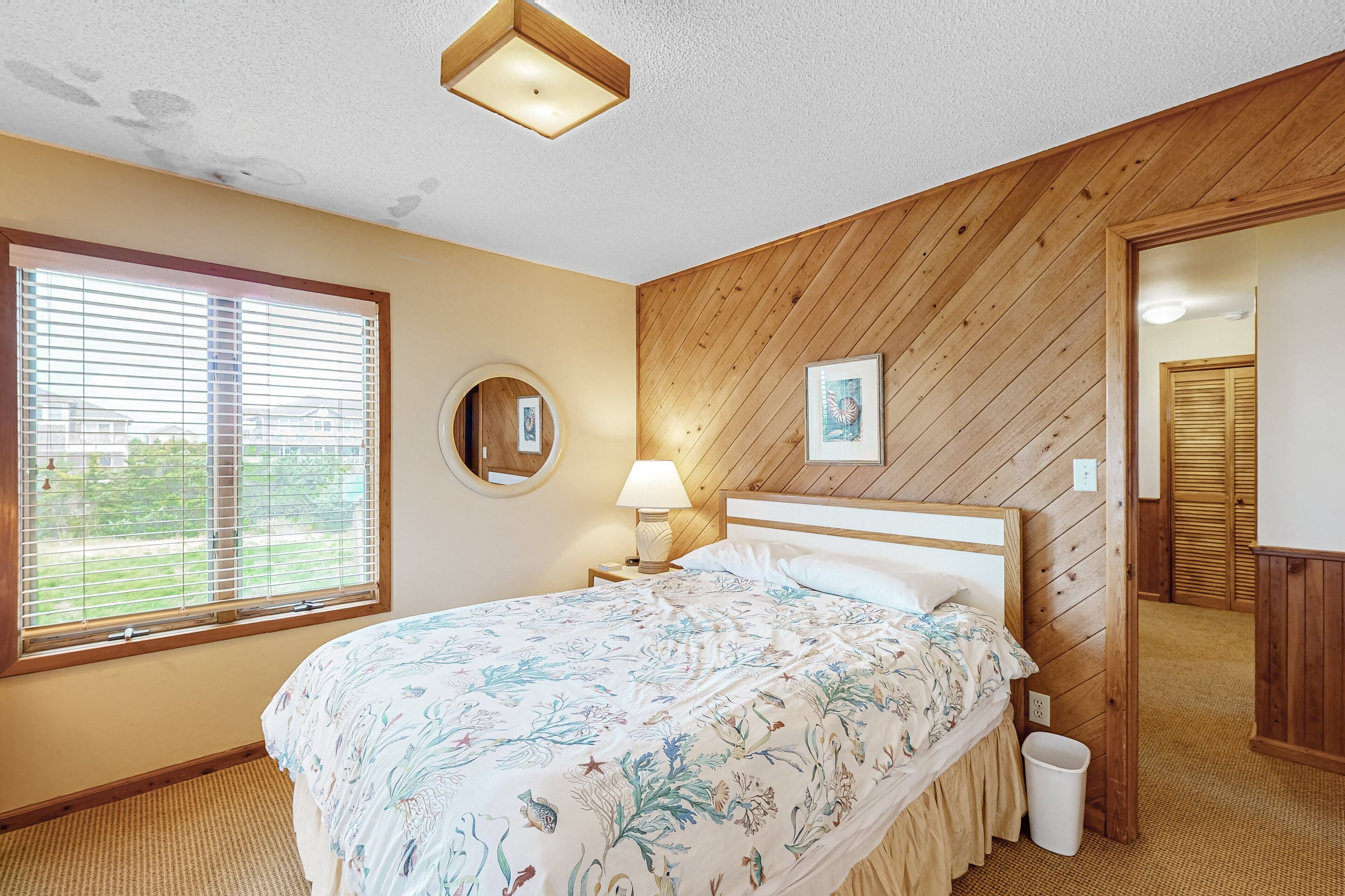 A bedroom with a wooden ceiling, a bed with floral bedding, a bedside lamp, a waste bin, and a window with blinds.