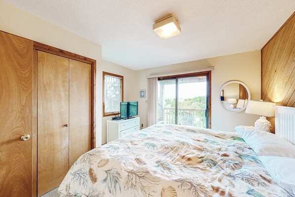 A bedroom with a large bed, a wooden wardrobe, a television on a dresser, a window with blinds, and a balcony view. - Charming, Oceanfront Home w/Free WiFi, Private Hot Tub, W/D, and Shared Pool (Avon)