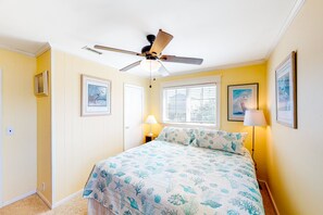 Une chambre à coucher avec un lit, un ventilateur de plafond, une fenêtre avec des stores et des tableaux encadrés accrochés aux murs.