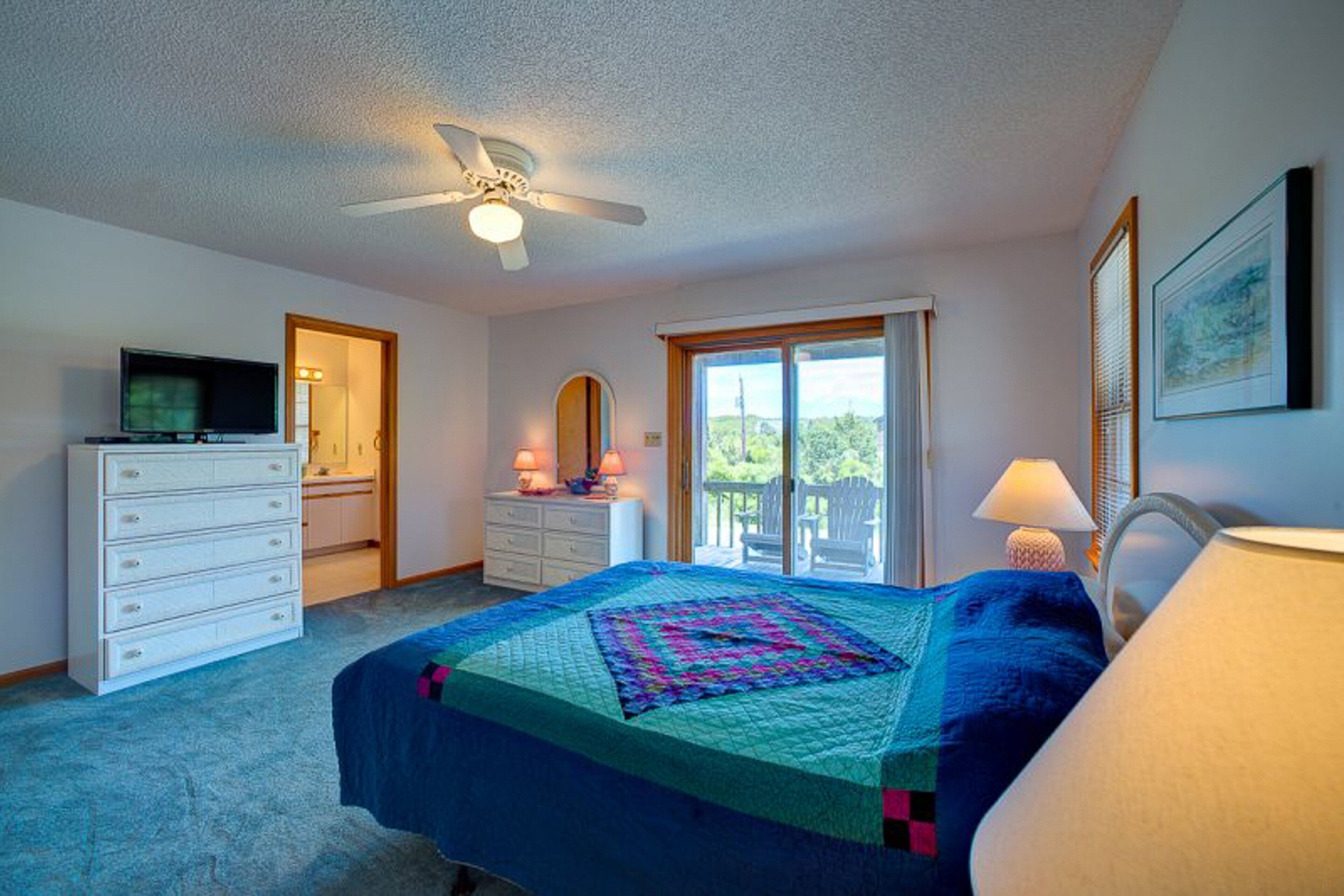 A bedroom with a bed, dresser, television, ceiling fan, and a window with a view.