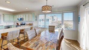 A modern kitchen with light green cabinets, a wooden dining table, wicker chairs, and a large window with blinds. - Sunset Park: Short Walk to the Beach, Great Views (Avon)